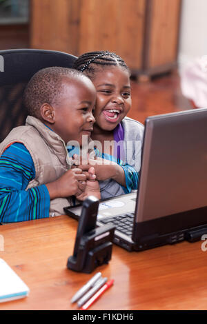 Young African sibling sot together at their father's desks, playing on laptop and laughing Stock Photo