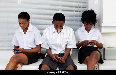 Three African teenagers sit together in a kitchen looking at their cellphones Stock Photo