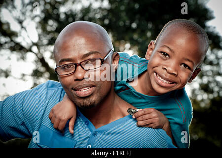 Young African boy and his father sit in a park, smiling at camera Stock Photo