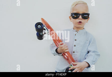 Adorable hipster little boy with a skateboard wearing borrowed sunglasses from his Mum or Dad standing against a wooden wall giv Stock Photo