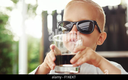 Cute hipster little boy in over sized sunglasses belonging to his mother or father sitting sipping a beverage in a glass on an o Stock Photo