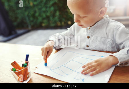 Small boy entertaining himself on a hot summer day standing at a table on an outdoor patio drawing with colored crayons on sheet Stock Photo
