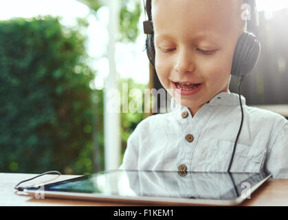 Adorable little boy listening to recorded music on his tablet computer using headphones with a delightful smile of contentment a Stock Photo