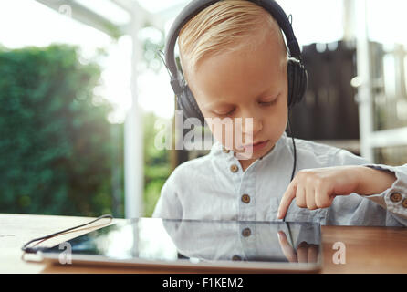 Pensive little boy selecting a new soundtrack from his music library on his tablet computer as he sits outdoors on a patio liste Stock Photo