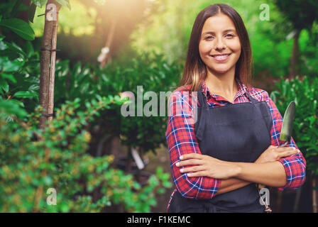 Smiling happy young woman enjoying her garden standing with folded arms against lush greenery with a trowel in her hand looking Stock Photo