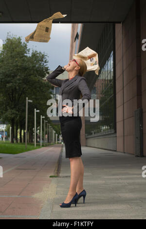 Young blonde business woman with a flying newspaper above her head in the city. Stock Photo