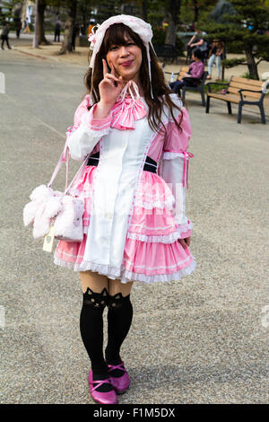 Young Japanese women in Osaka Park dressed in Goth-lolita pink and white maid costume, black kitten socks, posing for viewer. Eye-contact. Stock Photo
