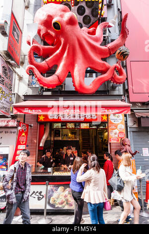 Japan, Osaka. Dotonbori. People outside Takoyaki restaurant, Dotonbori Kukuru Honten, with large red octopus sign above take away window. Stock Photo
