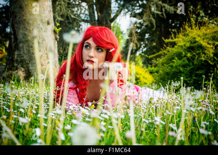 Young Woman with Bright Red Hair Lying Down in Forest Flower Field with Hand Resting on Chin Looking Dreamily Off Into Space Stock Photo