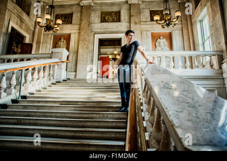 Young Handsome Man Standing on Stairs and Leaning Against Rail Inside an Architectural Building While Looking Into the Distance. Stock Photo