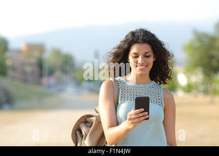 Front view of a happy girl walking and using a smart phone in a town Stock Photo