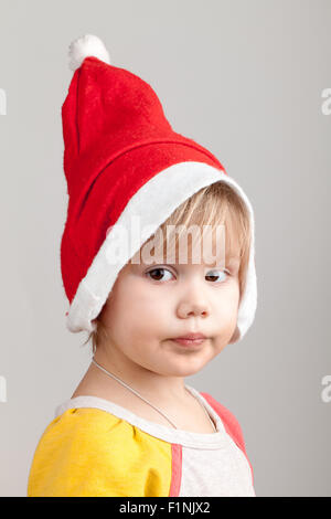 Cute Caucasian little girl in red Santa hat on gray background, closeup studio portrait Stock Photo