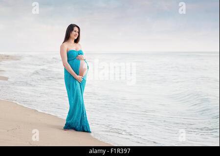 Pregnant woman wearing a turquoise blue maternity gown. She is smiling and looking out at the ocean. Stock Photo