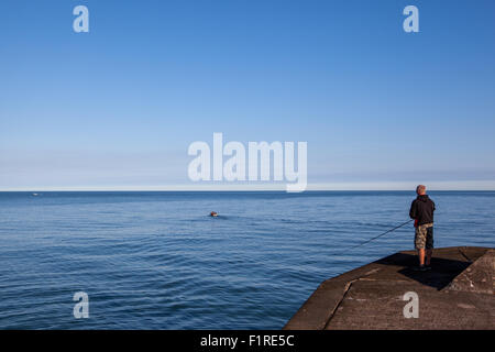 Small boat leaving shore as a man stands on the end of the jetty fishing Stock Photo