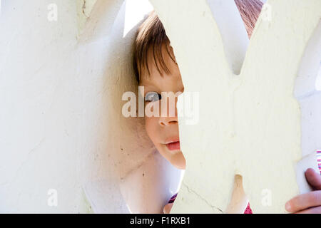 Caucasian child, boy, 3-4 year old, looking through gap in yellow stone wall directly at viewer. One eye viable as he peeks through. Eye-contact. Stock Photo