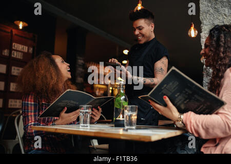 Cheerful two young women sitting at cafe holding menu card giving order to waiter. Young woman placing order to a waiter at rest Stock Photo