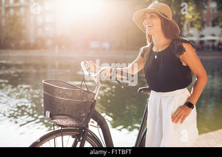 Portrait of beautiful young woman wearing a hat with a bicycle walking along a pond. Happy woman with a bike at the park looking Stock Photo