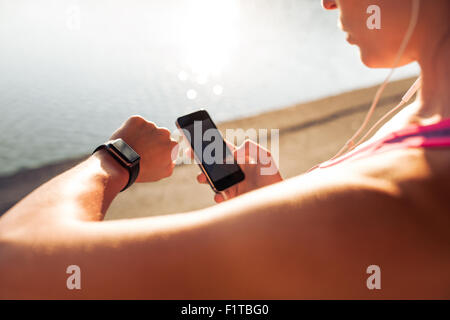 Sportswoman looking at smartwatch and holding smart phone in her other hand, outdoors. Fitness female setting up her smartwatch