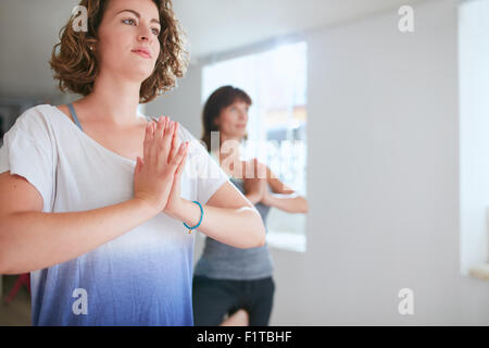 Portrait of young woman with her fitness trainer in background doing yoga exercising. Performing tree pose with hands clasped. V Stock Photo