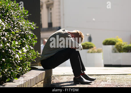Man sat on low wall with his head buried in his hands in Bedford, Bedfordshire, England Stock Photo