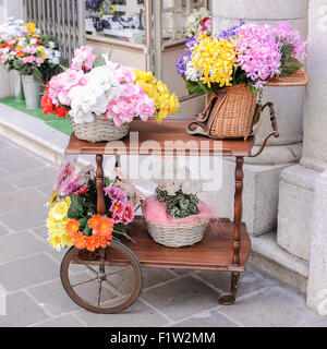 A cart with over wicker baskets and an old wicker fishing basket full of flowers for sale Stock Photo