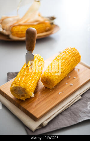 Ready, set, eat... Two rich, yellow corncobs are sitting on a wooden board, ready to be eaten. In one, a corn cob skewer is planted, making it nice and easy to eat. Stock Photo