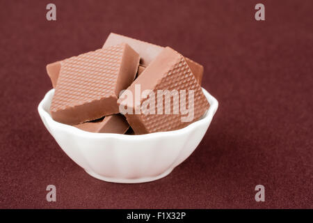 Chocolate squares in a white bowl on maroon background. Stock Photo