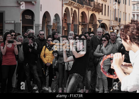 Focus on street musician playing the trumpet watched by a crowd with girl playing the tambourine in foreground Padova Italy Stock Photo