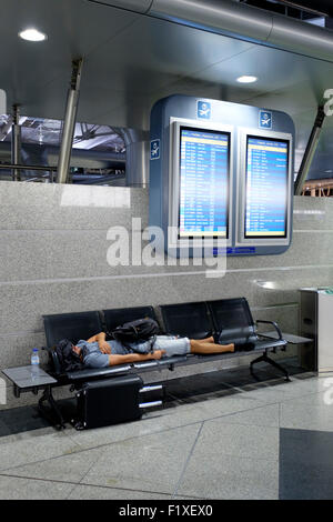 Passenger sleeping on a bench beneath two flight schedule boards at the Sá Carneiro airport terminal in Porto, Portugal Stock Photo