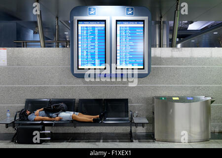 Passenger sleeping on a bench beneath two flight schedule boards at the Sá Carneiro airport terminal in Porto, Portugal Stock Photo