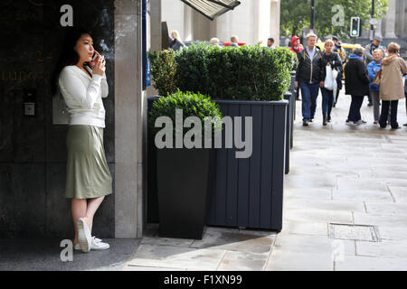 London 2015: An office worker takes a cigarette break outside her office building which fronts onto a busy city street. She using her mobile Stock Photo