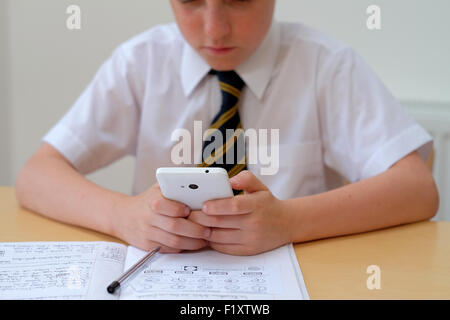 School pupil distracted by using a mobile phone (texting) whilst sat at his desk with his course work home work Stock Photo