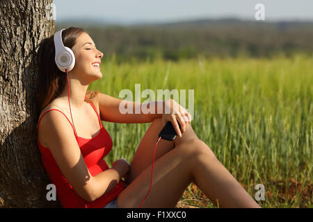 Casual girl listening to the music sitting and relaxed in a green field at sunset wearing a red shirt Stock Photo