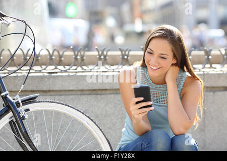 Happy girl watching media in a smart phone sitting in the street beside her bicycle Stock Photo