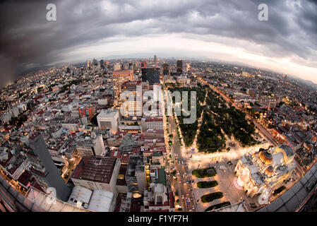 Mexico City Aerial Fisheye Alameda Central Stormy Sky | MEXICO CITY, Mexico — A fisheye aerial view from the Torre Latinoamericana's 44th-floor observation deck takes in the Palacio de Bellas Artes at lower right and Alameda Central spreading across the frame under a stormy evening sky. Laid out in the late sixteenth century, Alameda Central is the oldest formal public park in the Americas, its tree canopy marking the western edge of the Centro Histórico, the UNESCO World Heritage district built on the drained lakebed of the Aztec capital Tenochtitlan. Stock Photo