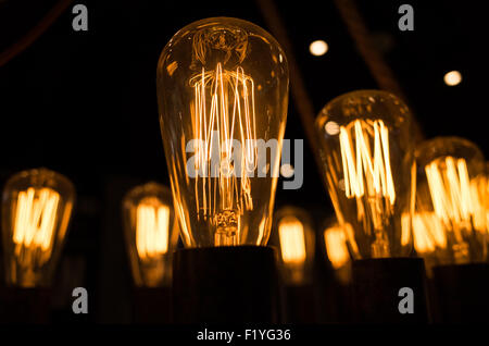 Vintage Edison Style Light Bulbs Intricate Glowing Filaments Close Up | Close-up of vintage Edison-style light bulbs shows their intricate looped filaments glowing against a dark background. These squirrel-cage carbon or tungsten filaments, a hallmark of late 19th-century incandescent design, produce a warm amber light prized today for decorative rather than high-output lighting. Multiple bulbs recede into soft focus, emphasizing the sharply detailed filament pattern in the foreground bulb. Stock Photo