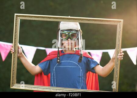 Portrait of boy looking through picture frame wearing cape, goggles and flying hat Stock Photo