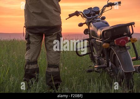 Mid adult man, standing in field, next to motorbike, at sunset, rear view, low section Stock Photo