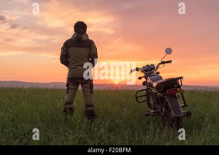 Mid adult man, standing in field, next to motorbike, at sunset, rear view Stock Photo