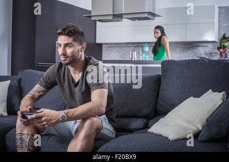 Young man on sofa using gaming control watched by angry girlfriend Stock Photo