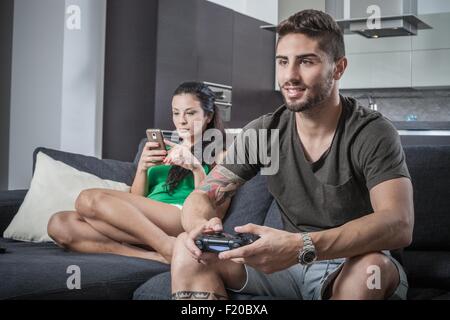 Young couple on sofa using smartphone and gaming control Stock Photo