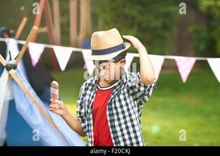 Boy eating ice lolly whilst adjusting trilby hat in garden Stock Photo