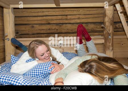 Two young women lying on bed in log cabin chatting Stock Photo