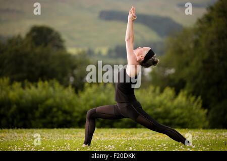 Mature woman practicing yoga warrior pose with arms raised in field Stock Photo