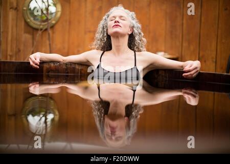 Mature woman meditating with eyes closed in hot tub at eco retreat Stock Photo