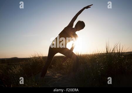 Rear view of mid adult woman practicing yoga in silhouetted  long grass at sunset Stock Photo