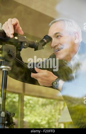 Senior man at home, using telescope through window Stock Photo