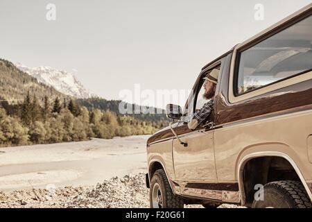 Mid adult man in pickup truck, leaning out of window looking at mountain range, Wallgau, Bavaria, germany Stock Photo