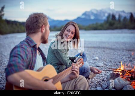 Young couple sitting by campfire playing guitar, Wallgau, Bavaria, Germany