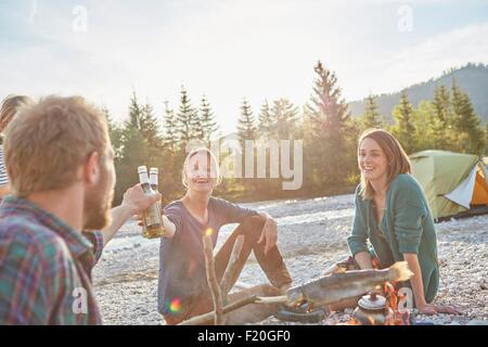 Adults sitting around campfire making a toast with beer bottles Stock Photo
